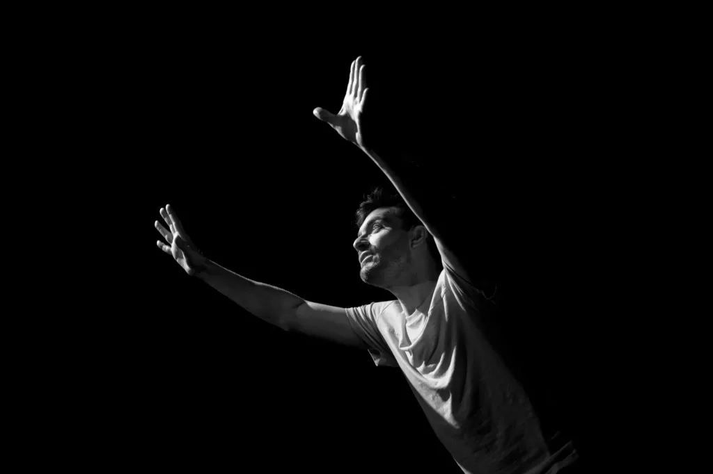 Damien Saugeon lève les mains et le regard vers le ciel sur une scène sombre dans le spectacle Certains regardent les étoiles